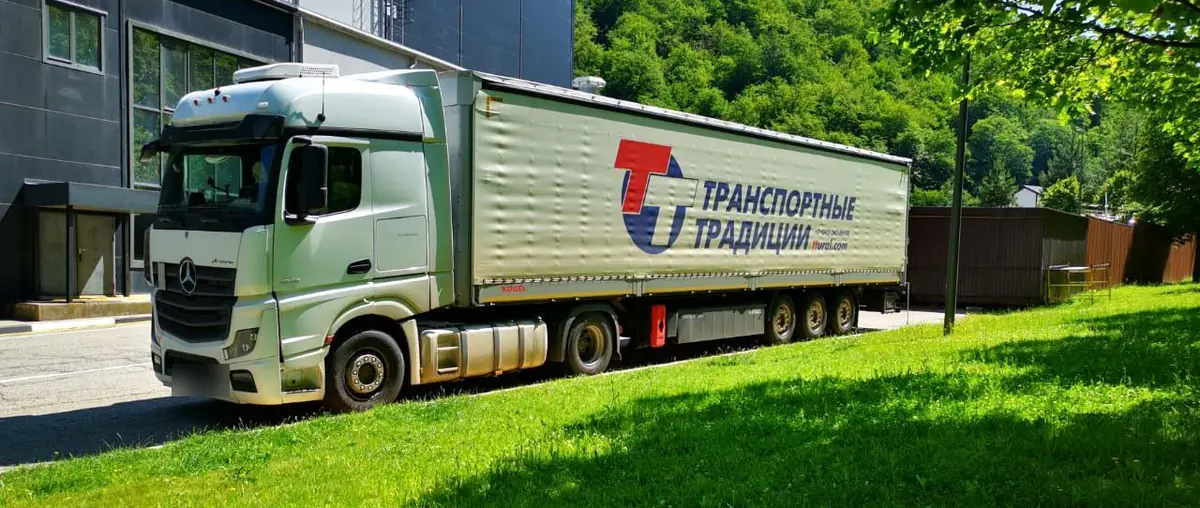 Автопоезд компании в фирменной оклейке у производственного корпуса, на заднем плане лесистые горы
